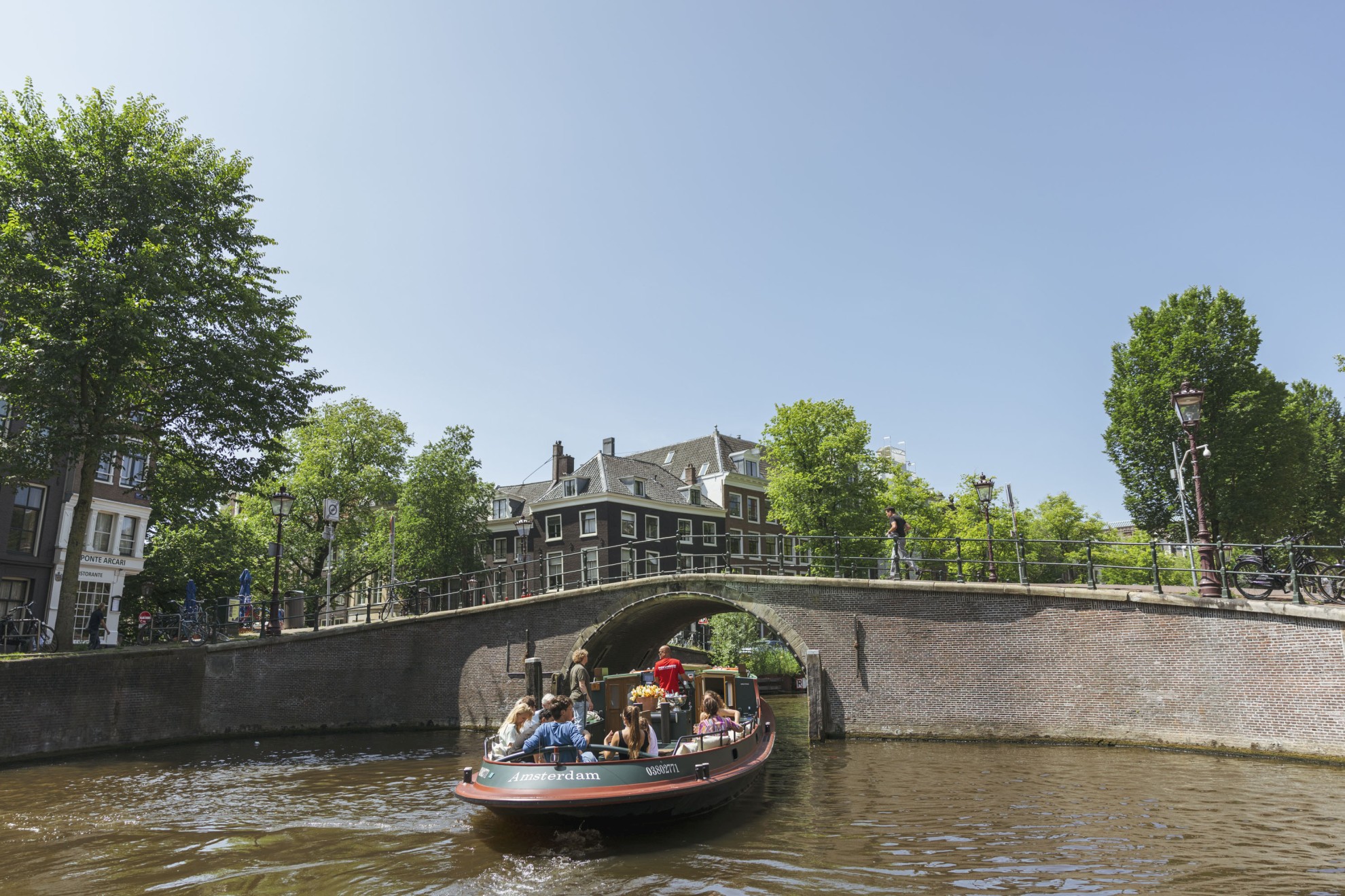 Ein Boot voller Passagiere fährt unter einer Steinbrücke auf den Amsterdamer Grachten hindurch, umgeben von historischen Gebäuden und viel Grün. Ein Boot voller Passagiere fährt unter einer Steinbrücke auf den Amsterdamer Grachten hindurch, umgeben von historischen Gebäuden und viel Grün.