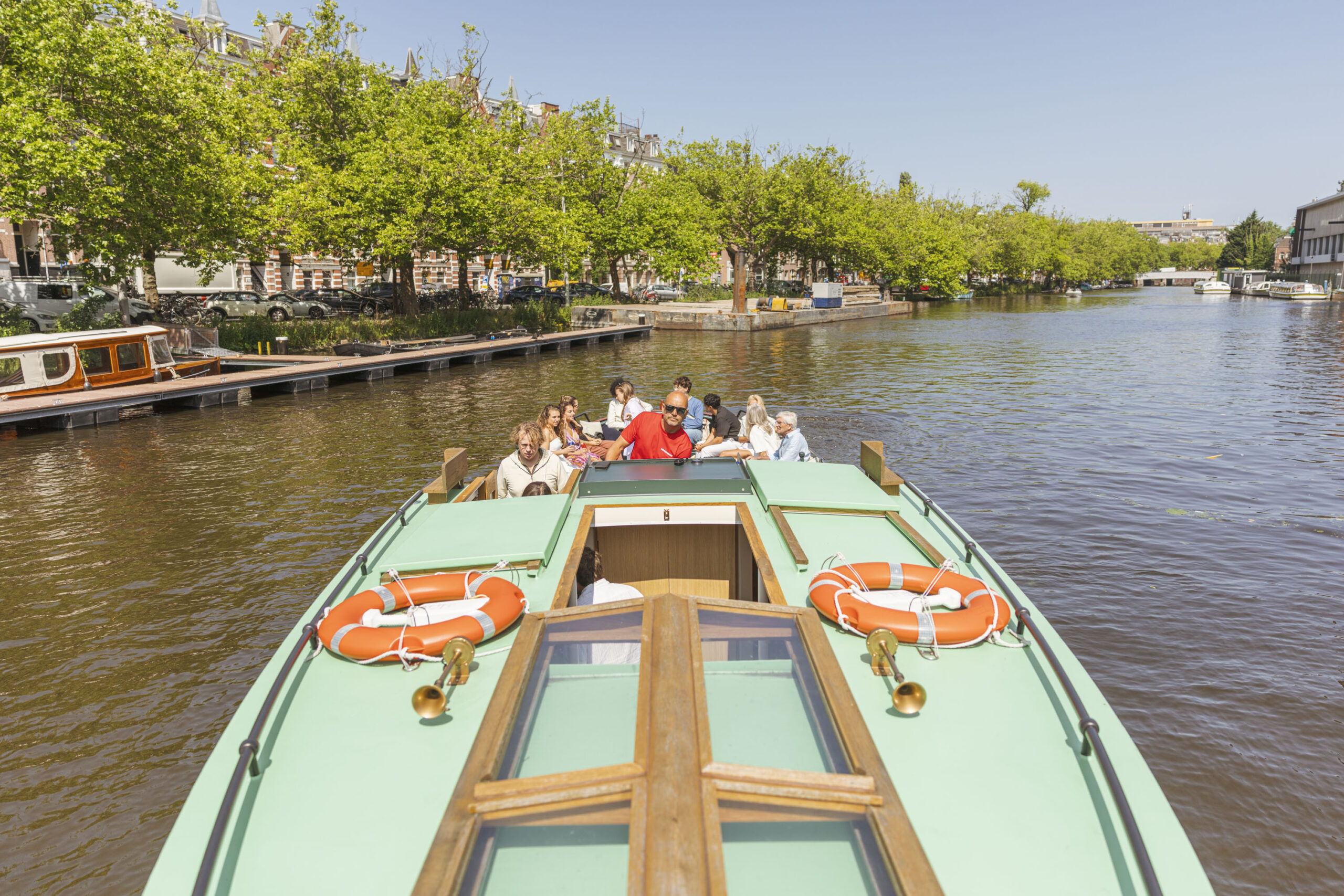 A group of people sit relaxed on a classic canal boat in Amsterdam, as the skipper steers the vessel through the canal on a sunny day. A group of people sit relaxed on a classic canal boat in Amsterdam, as the skipper steers the vessel through the canal on a sunny day.
