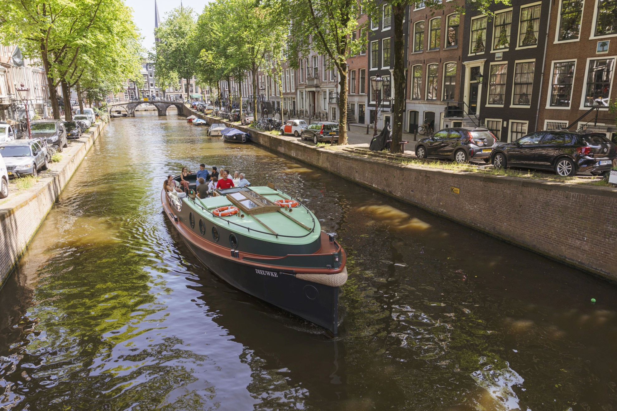 Boaty.nl - Rondvaartboot op Amsterdamse gracht Eine Gruppe von Menschen fährt in einem klassischen Boot durch einen Amsterdamer Kanal, umgeben von Bäumen, geparkten Autos und historischen Kanalhäusern.