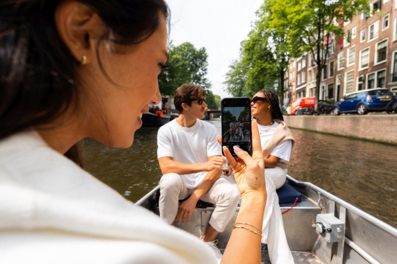 Freunde machen ein Foto während einer entspannten Bootsfahrt durch einen Amsterdamer Kanal, umgeben von historischen Grachtenhäusern und grünen Bäumen.