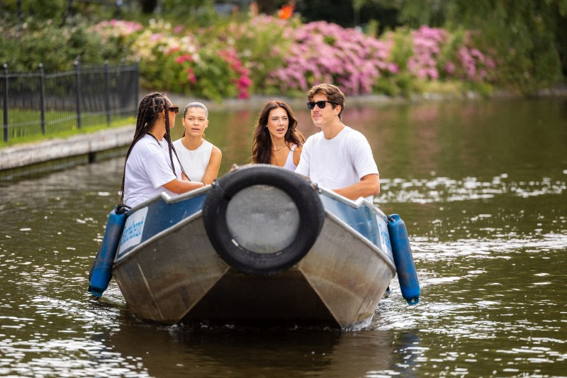 Vier Freunde segeln gemeinsam in einem kleinen Boot auf einem ruhigen Kanal, umgeben von Grün und blühenden Büschen.