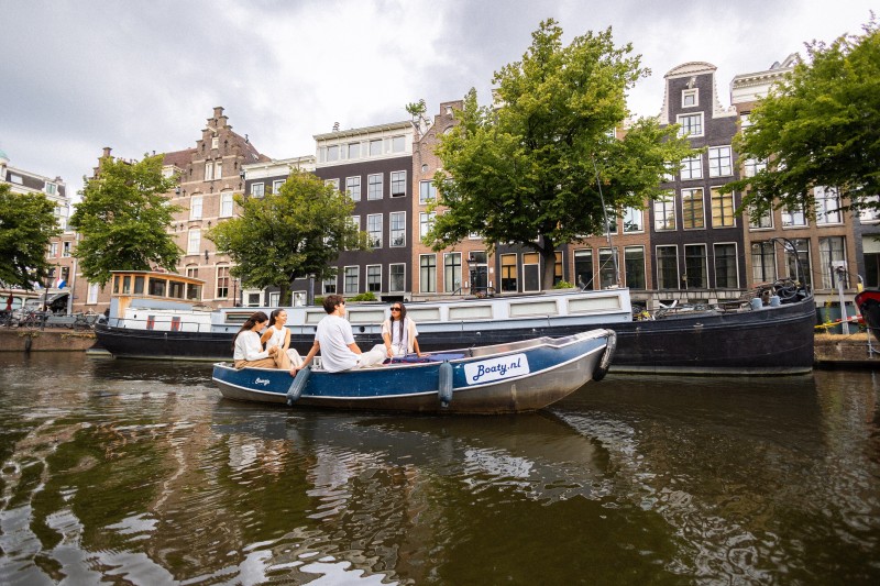 Eine Gruppe von Freunden segelt in einem kleinen Mietboot durch einen Amsterdamer Kanal, vorbei an historischen Grachtenhäusern und Hausbooten.