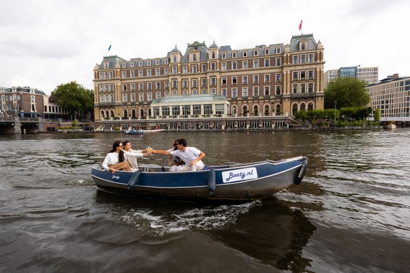Freunde stoßen bei einer Bootsfahrt auf dem Wasser mit Getränken an, im Hintergrund ist ein historisches Gebäude entlang des Amsterdamer Kanals zu sehen.